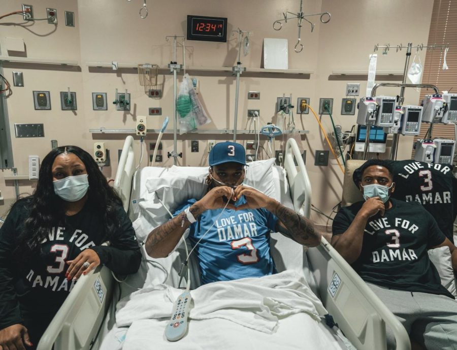 Hamlin poses from his hospital bed surrounded by families, displaying a heart sign in solidarity for the next Bills game after injury.